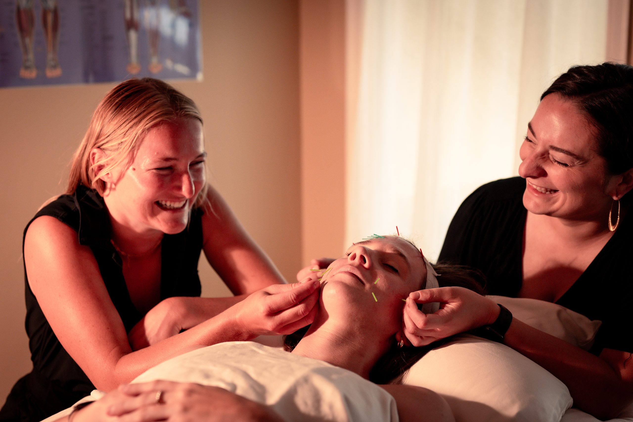 Water Tiger Acupuncturists Jamie and Dana smile as they place facial acupuncture needles in patient, who is smiling as well.